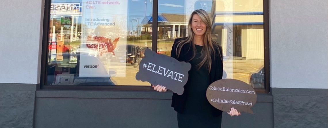 Cellular Sales team member standing in front of store with 2 signs that read #elevate and #cellularsalesproud