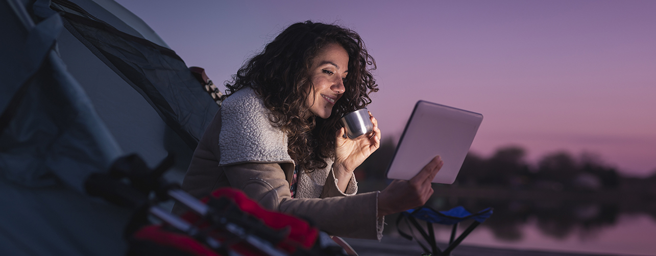 woman sipping drink from cup in a tent while streaming a show on her iPad