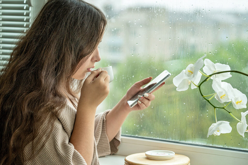 A woman with long hair sits by a window, sips from a cup, and looks at her phone as it rains outside. 