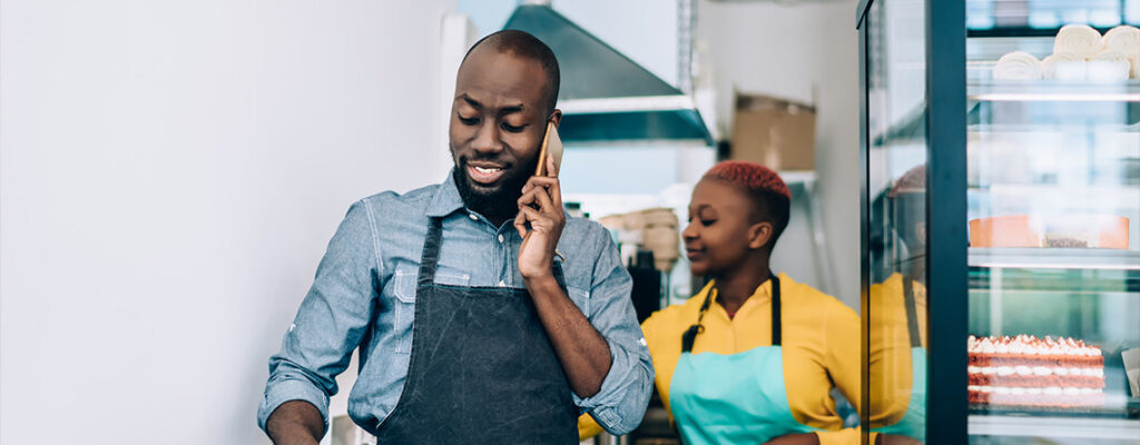 A man wearing an apron talks on the phone next to a bakery case displaying cakes.