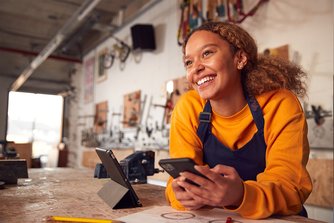 A curly-haired woman in a gold sweatshirt leans against a counter smiling and holding her phone.