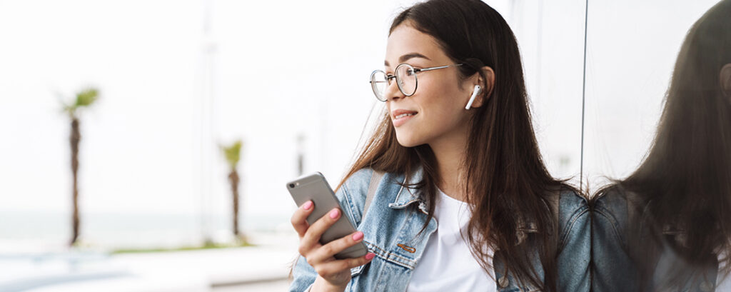 A woman with long dark hair and glasses listens to music on her smartphone while wearing AirPods.