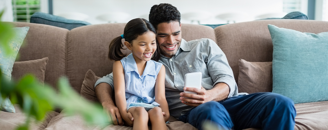 A man and small girl are smiling at a phone together, seated on a couch.