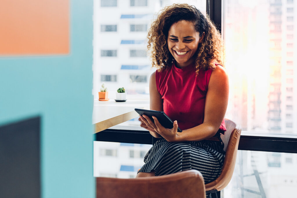 A young woman with brown skin and curly hair is holding a smartphone and smiling at it. She wear a pink sleeveless top and is sitting in a brown chair in front of a window.