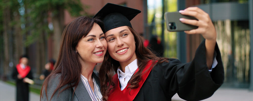 A mother and daughter pose for a selfie. The daughter is wearing a graduation gown and mortarboard, and is holding a smartphone.