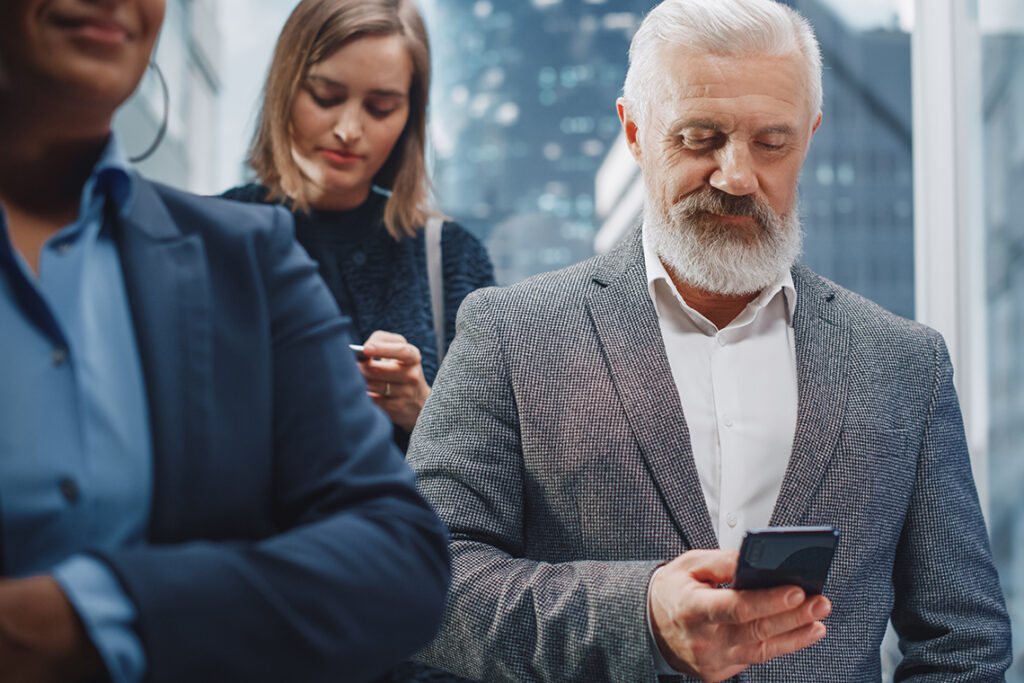 An older man in a gray suit smiles down at his phone in a room with other people.