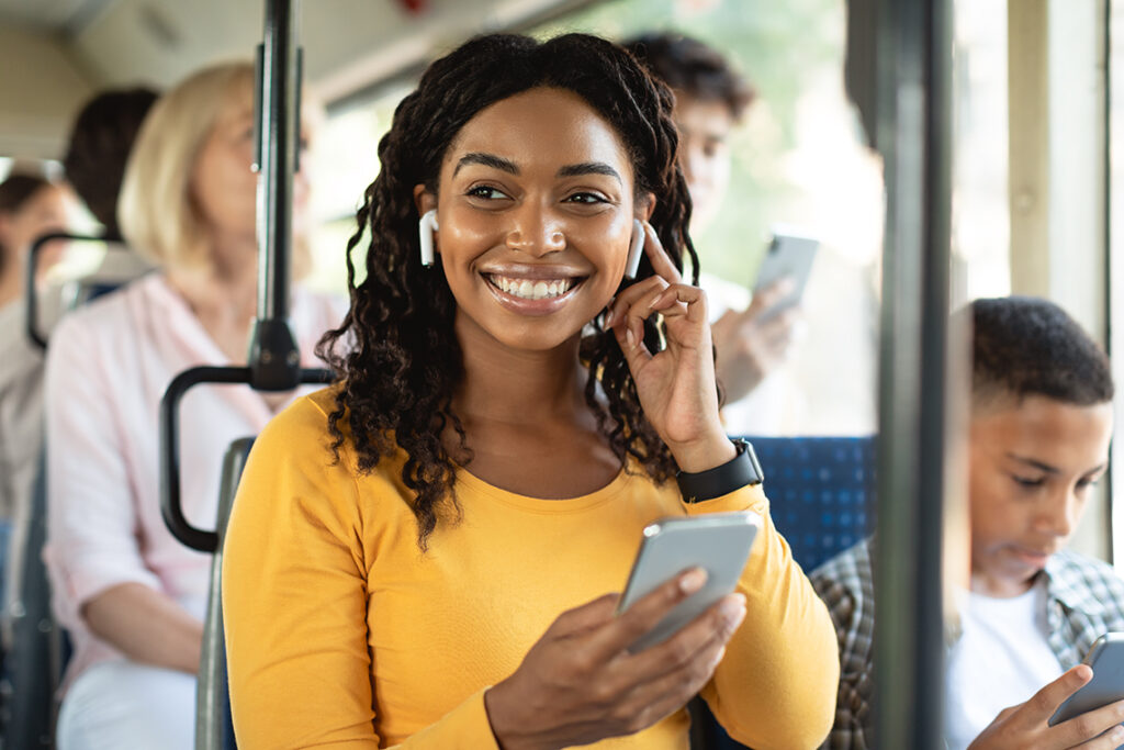 A young Black woman in a yellow top is sitting on a bus with other passengers, listening to music with her AirPods and smartphone, and smiling.