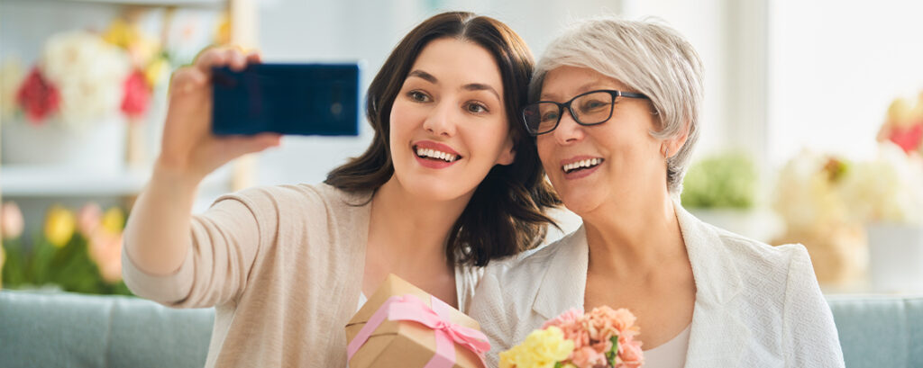 A young woman with dark hair holds a phone to take a selfie with an older woman in glasses.