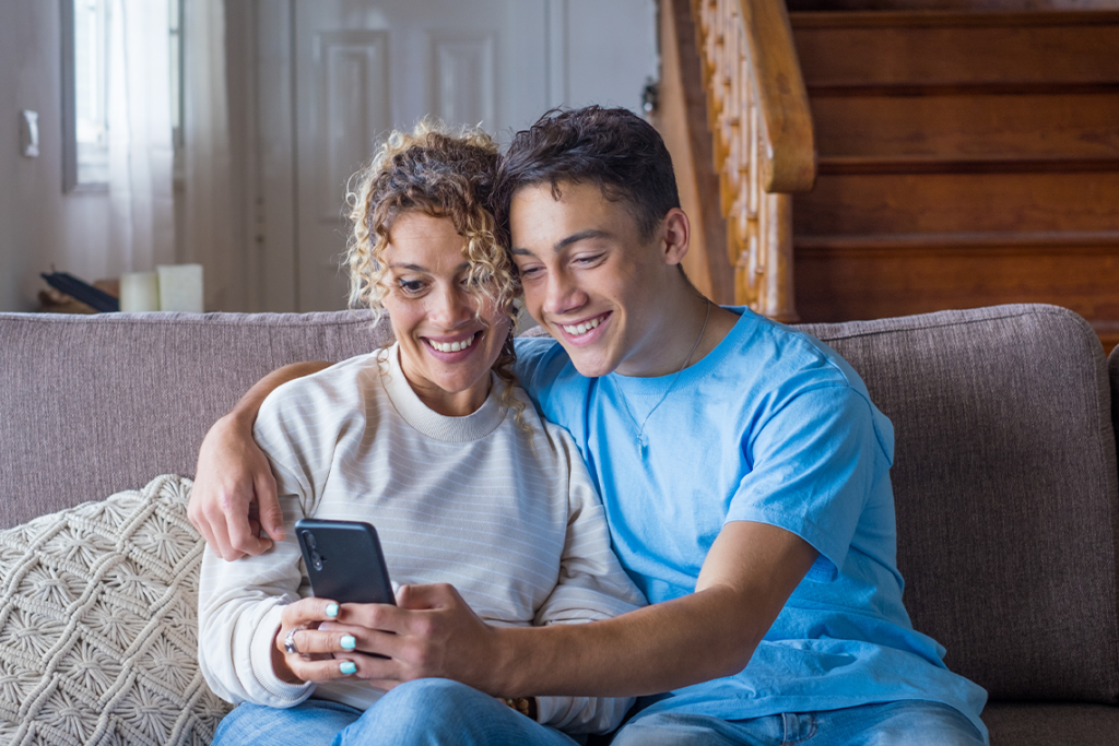 A husband and wife sit together on a couch, looking down at a phone and smiling.