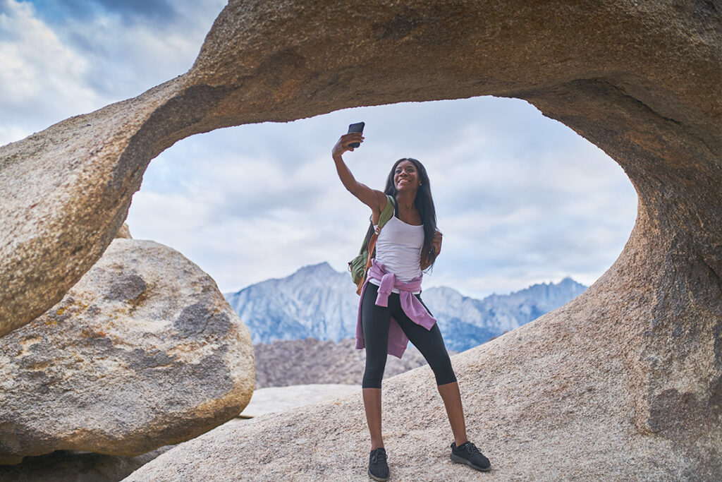A young Black woman stands in a rocky landscape. She is holding her phone in the air and smiling for a selfie.