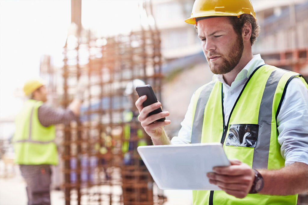 A bearded man in a high-visibility vest and hard hat stands in a construction site. He is holding a tablet and looking seriously at his phone.
