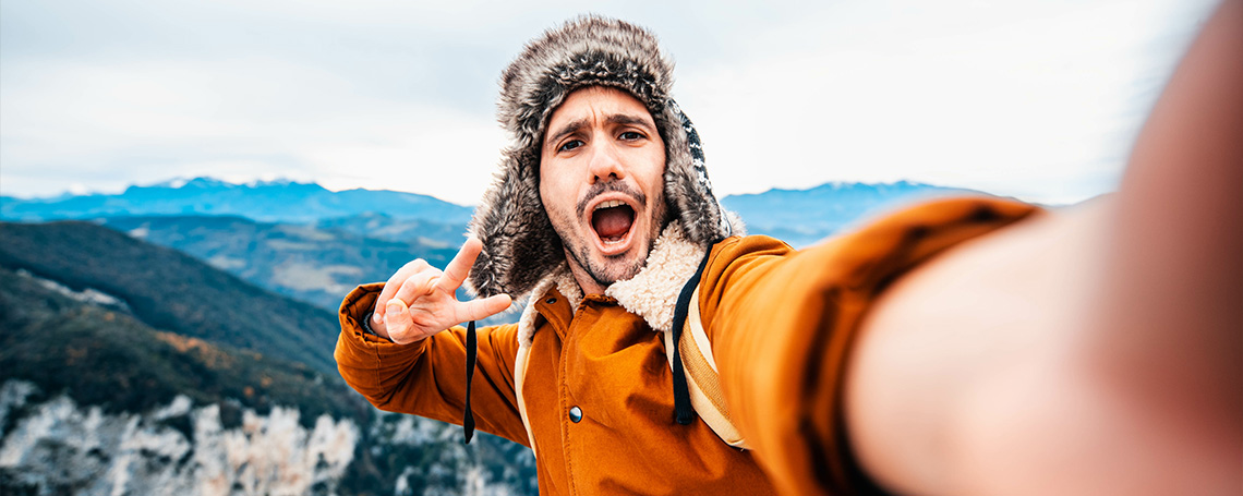 A man in an orange parka and fuzzy hat poses for a selfie, holds up a peace sign.