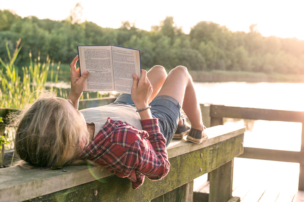 A child reads a book while lying on a wooden bench next to a lake during sunset.