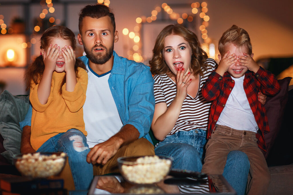 A family of four sits on the couch, eating popcorn and watching a movie. The children cover their eyes as they sit on their parents' laps.