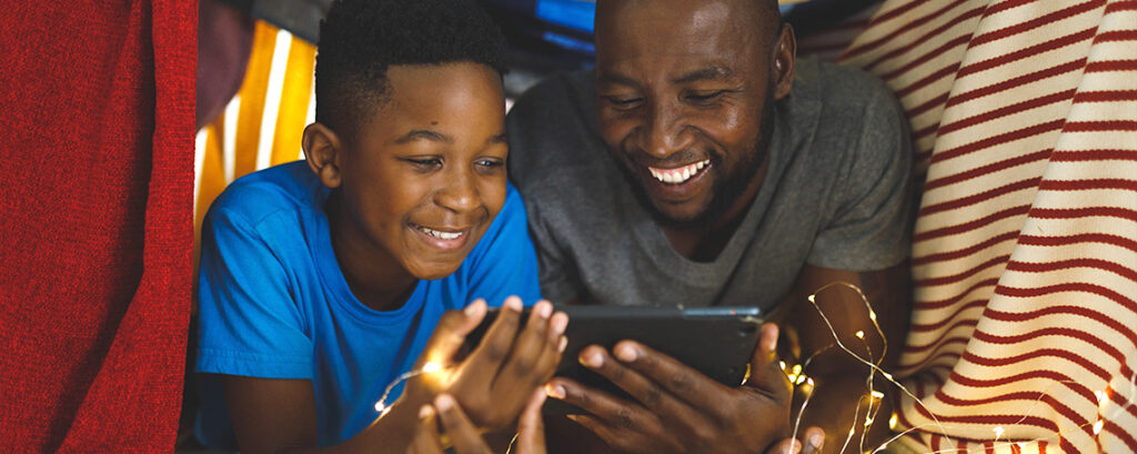 A Black man and his son lay in a blanket fort, laughing and smiling while looking at their smartphones.