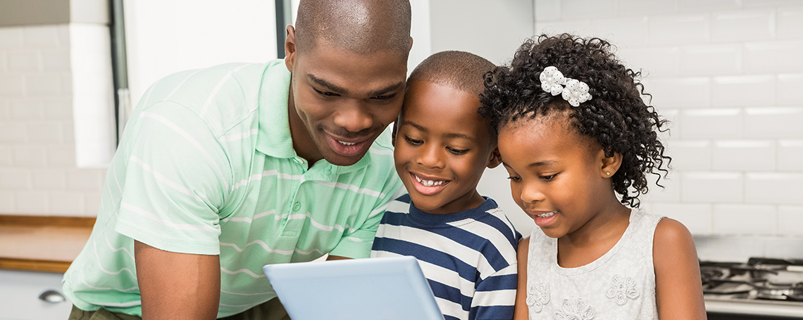 A father and two young children sit together, smiling and watching something on a tablet.
