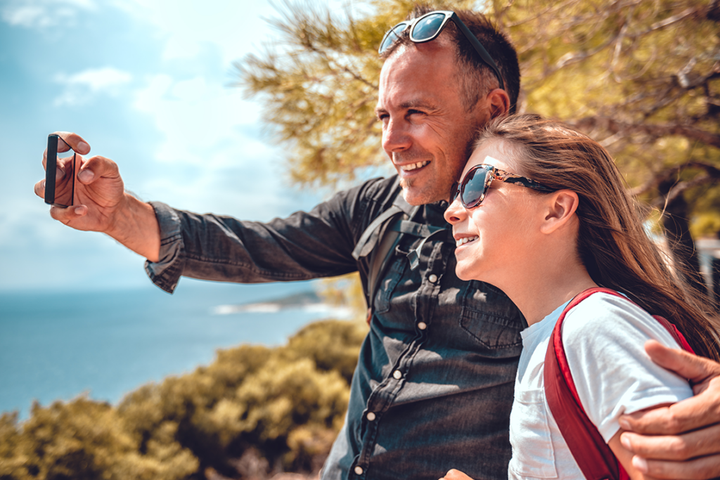 A man and his daughter pose for a smartphone selfie while hiking near the sea. The man is holding the phone and has his arm around the daughter.