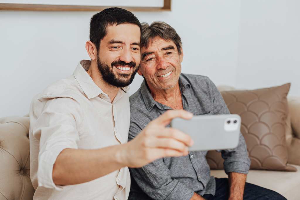A young bearded man holds a phone for a selfie with an older man. Both are smiling at the phone.