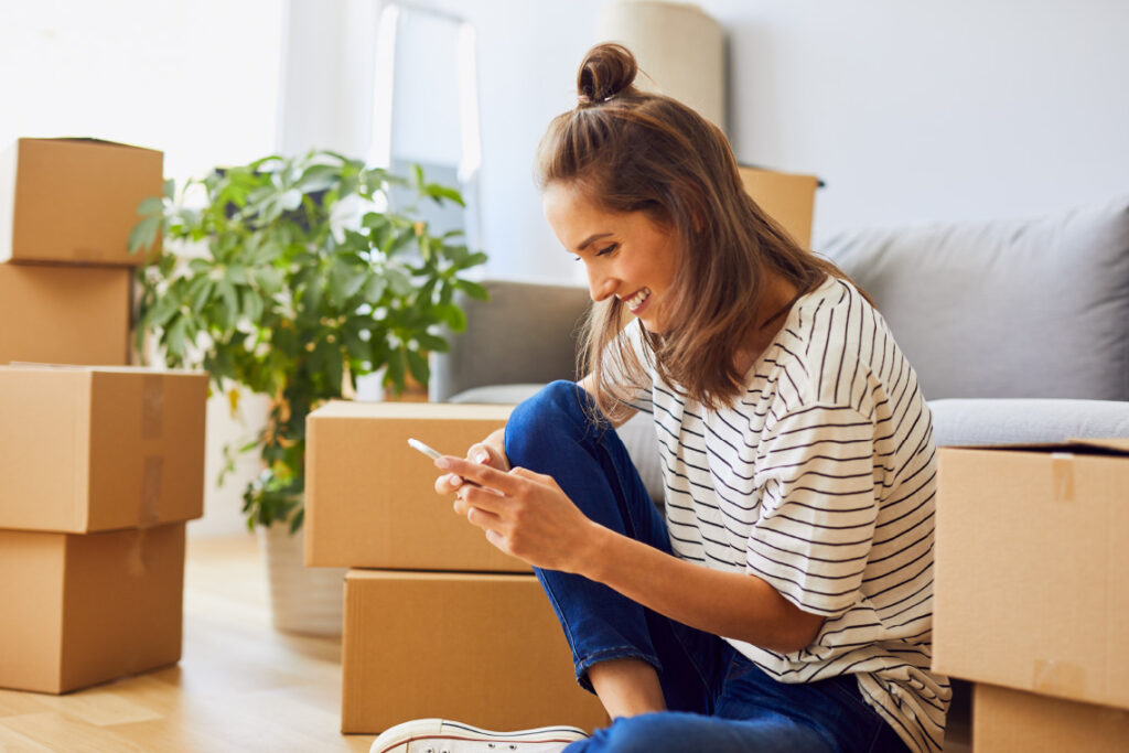 A woman sits in a sunny room, smiling at her phone. She is surrounded by moving boxes.