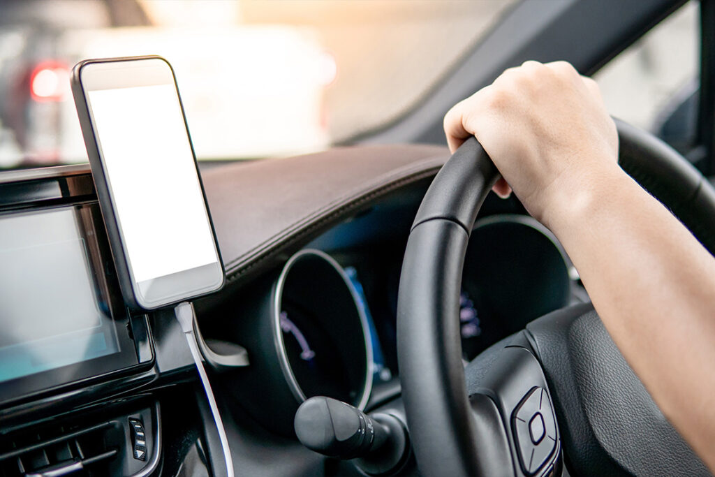 A phone sits in a dashboard mount, attached to a charging cord. In the background, a forearm and hand are visible, holding a car steering wheel.