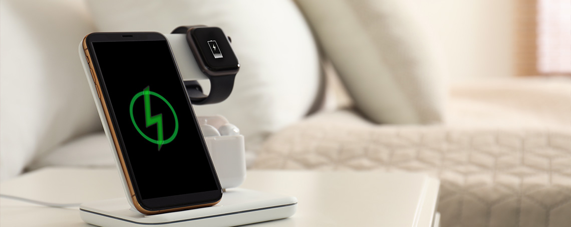 A black smartphone sits on a charger on top of a bedside table. The screen shows a charging icon.