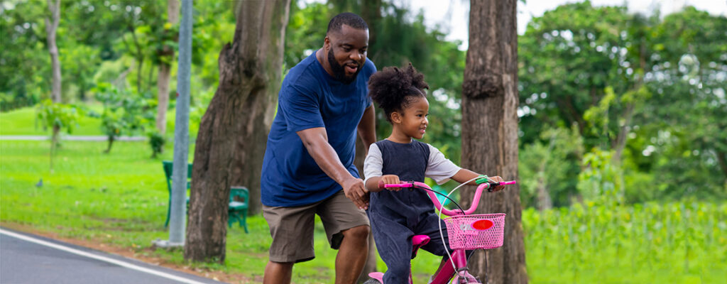 A Black father pushes his young daughter on a pink bike as she learns to ride.