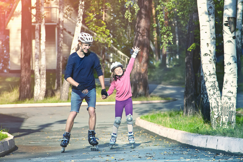A parent and young child rollerblade together through a neighborhood, smiling and holding hands.