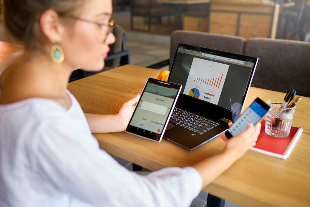 A young woman in a white shirt and glasses sits at a desk, looking at info on a tablet, smartphone, and laptop screen simultaneously.