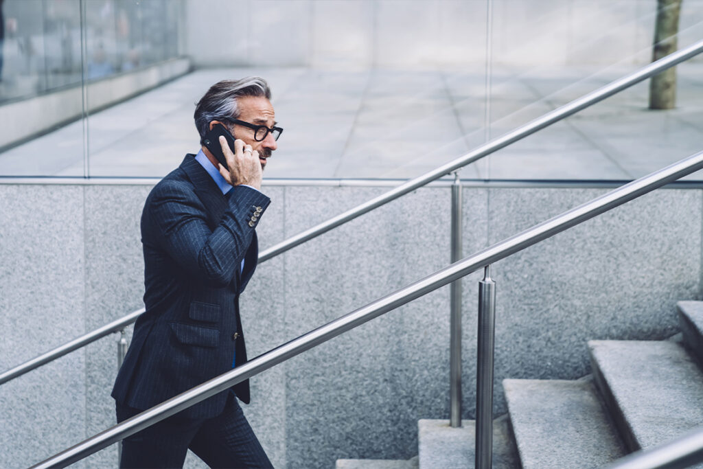A middle aged man wearing a dark blue suit and glasses walks up concrete stairs while talking on this phone.