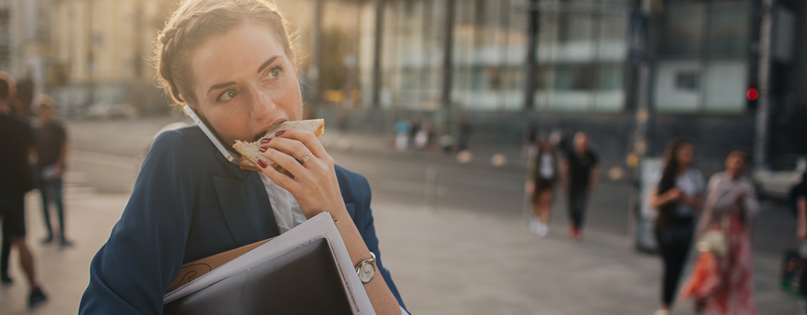 A woman with braided brown hair eats a sandwich and holds a phone between her ear and shoulder while rushing to work.