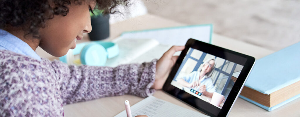 A young child with brown skin and curly hair watches a school lesson on their tablet.