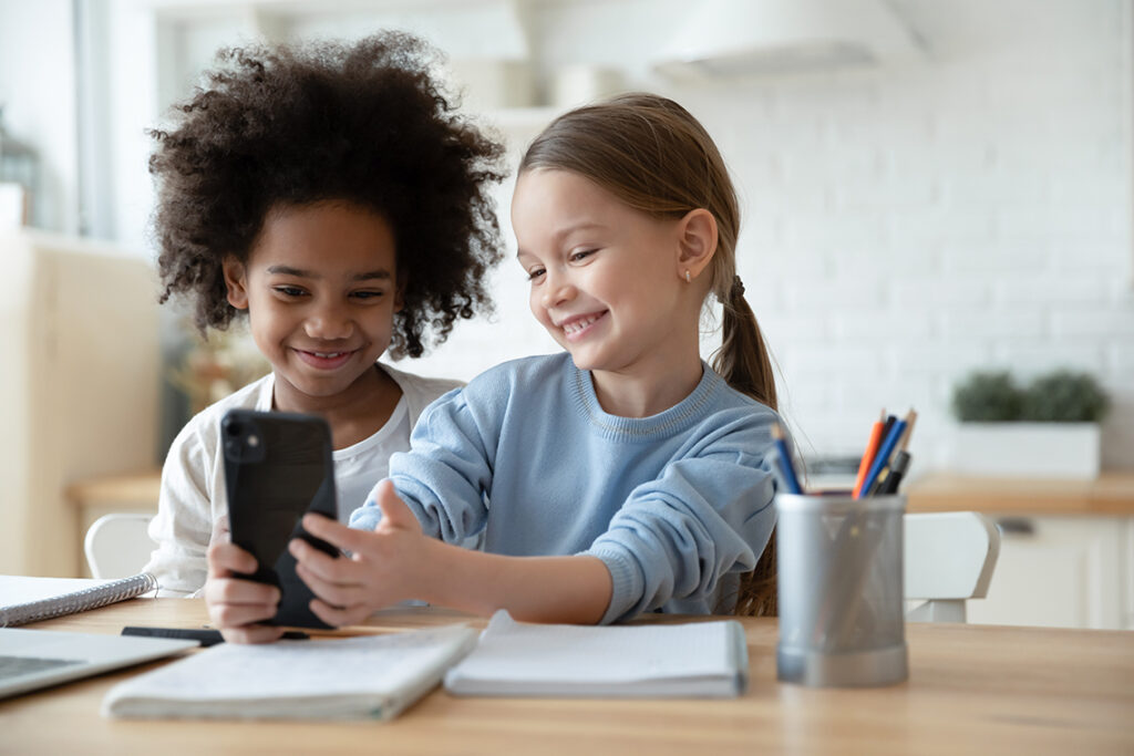 Two elementary-aged schoolchildren sit next to each other, smiling and use a smartphone while working together.