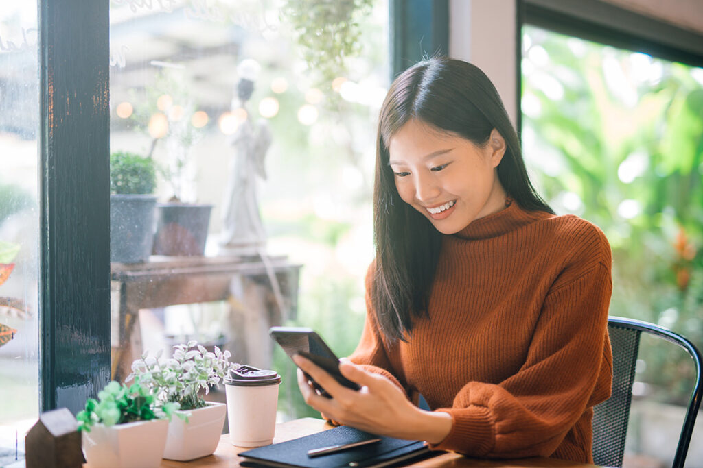 An Asian woman sits in a cafe, next to a window. She's holding her smartphone and smiling at the screen.