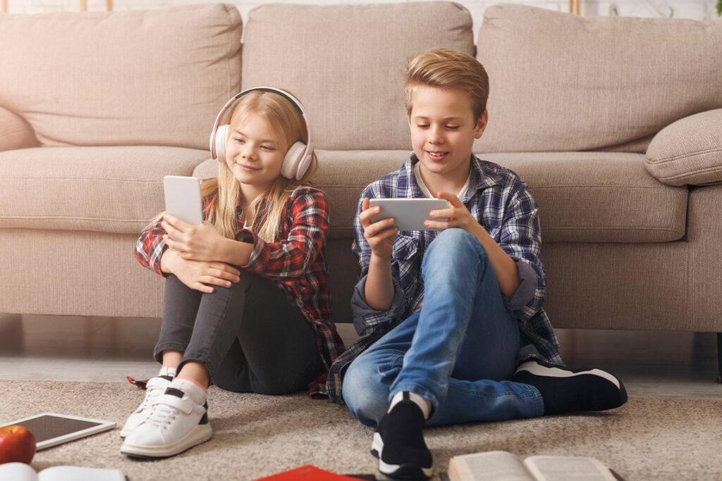 Two children, a boy and a girl, sit on the floor in front of a beige sofa. They're holding smartphones, playing games and listening to music.