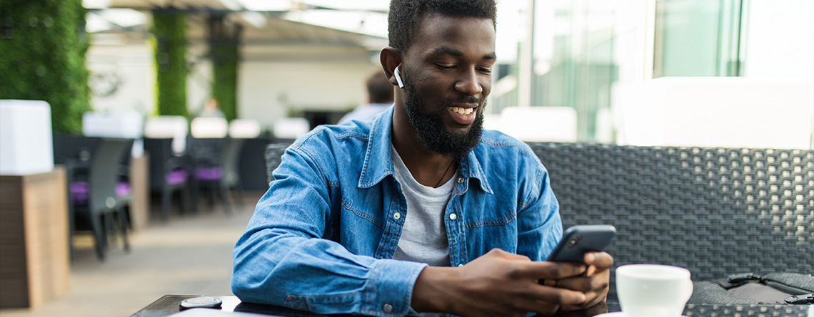 A young Black man wearing a denim shirt sits at a table. He's wearing a Bluetooth ear bud and smiling down at his phone.