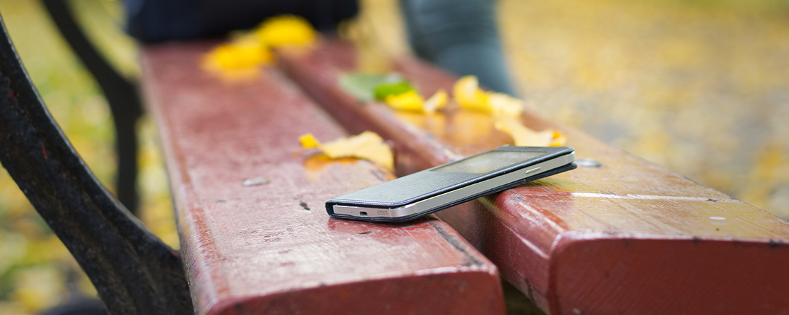 A phone is left unattended on a park bench, with yellow leaves and a person's legs in the background.