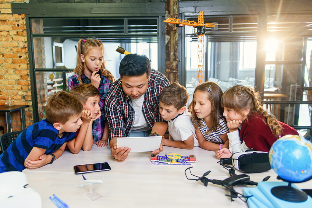 A male teacher sits with six students at a table, on which are a smartphone, a VR headset, and a drone. They are all looking at a tablet that the teacher is holding.