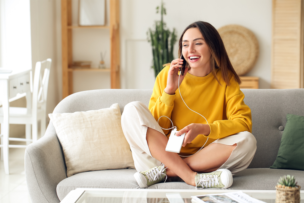 A woman in a yellow sweatshirt sits on a sofa, talking on her smartphone, which she is also charging with a portable charger.