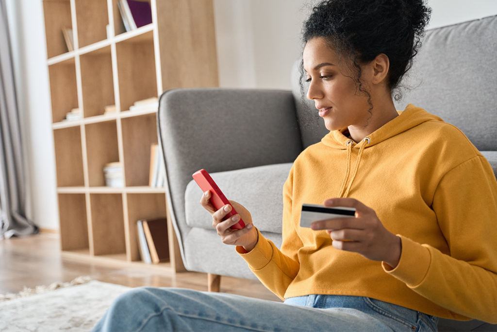 A woman with curly hair and wearing a yellow hoodie sits in front of her sofa, holding a credit card in one hand and her smartphone in the other.