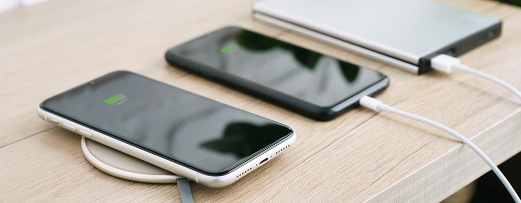 Two smartphones sit charging on a table, one wirelessly and the other with a portable charger.