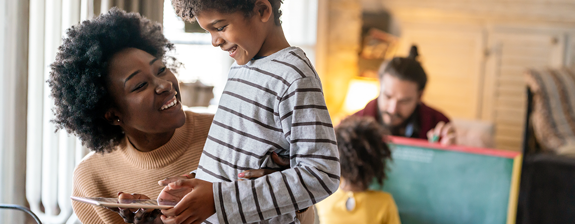 A Black woman with natural hair, is kneeling and smiling up at her student, a young boy in a striped shirt. In the background, a white man uses a chalkboard on an easel with another child.