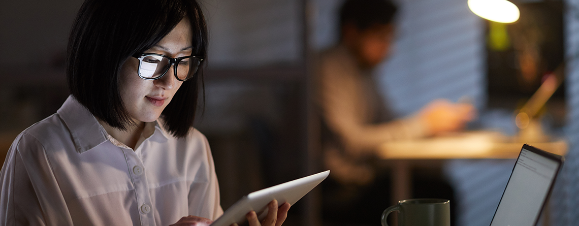 An Asian woman with glasses holds a tablet in her hands and stares down at the screen.