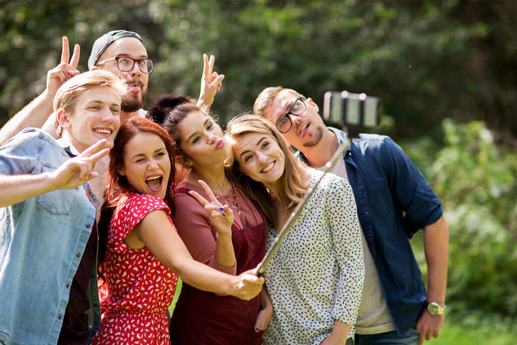 A family stands outside, making silly faces as they pose for a group selfie. A woman in front holds a smartphone on a selfie stick.