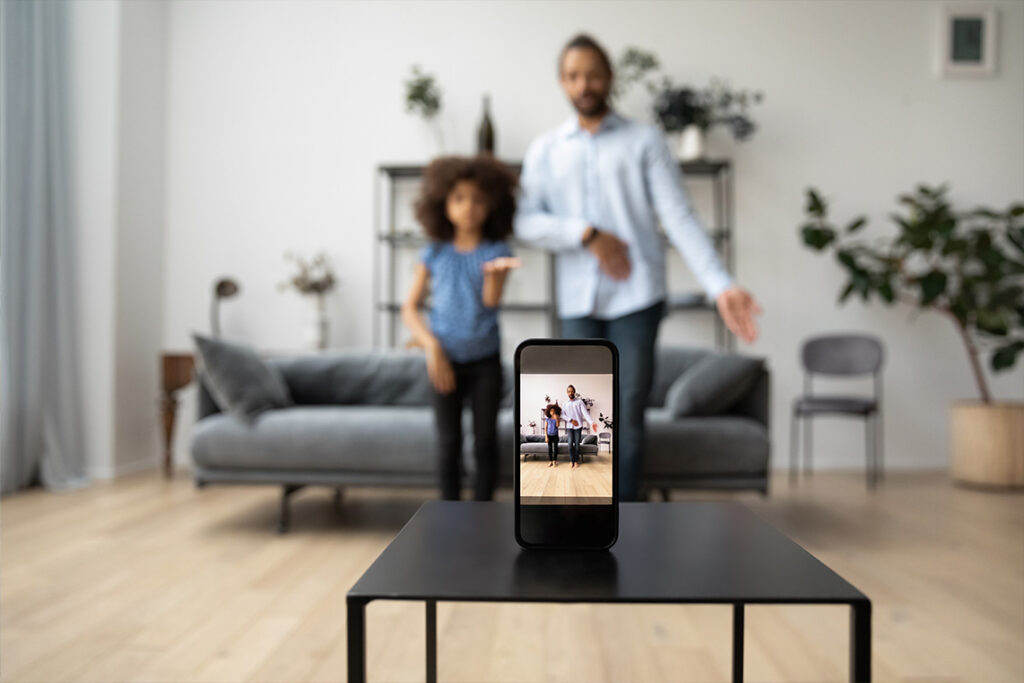 A father and daughter pose in their living room. A smartphone in the foreground is recording them.
