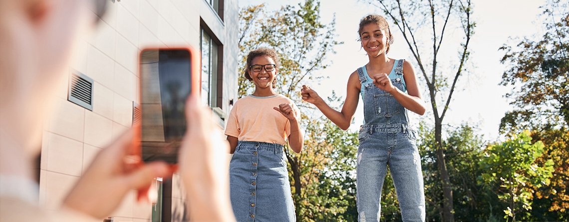 Two young girls dance and smile as their parent records a video on their smartphone.