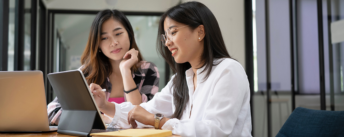 Two young women sit at a table, using a tablet and stylus. One woman is wearing a white blouse and the other is wearing a pink and black plaid shirt.