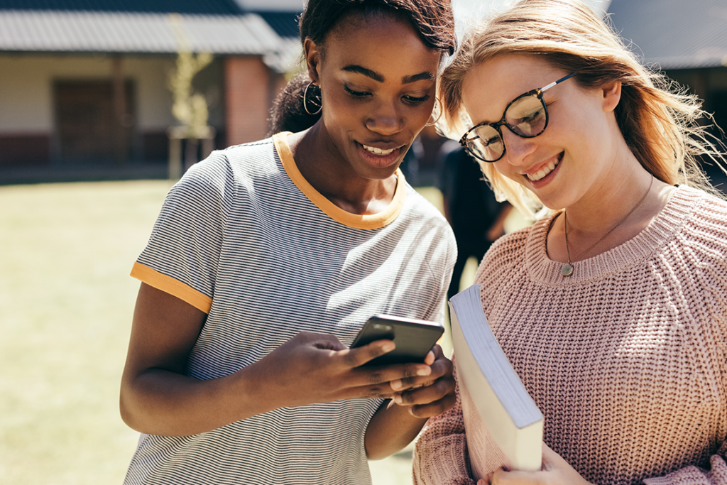 Two young women are standing on a college campus. One is holding a smartphone and they're both looking down at it.