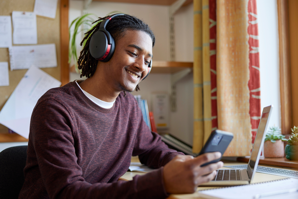 A young man in a knit sweater studies at his desk. He's smiling down at a smartphone and wearing wireless headphones.