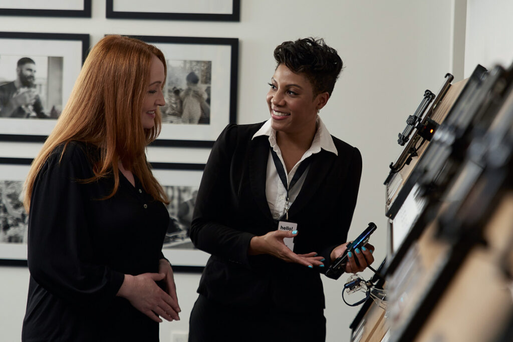 A sales rep in a dark suit jacket and white blouse helps a red haired female customer. They're standing next to a shelf full of wireless devices.