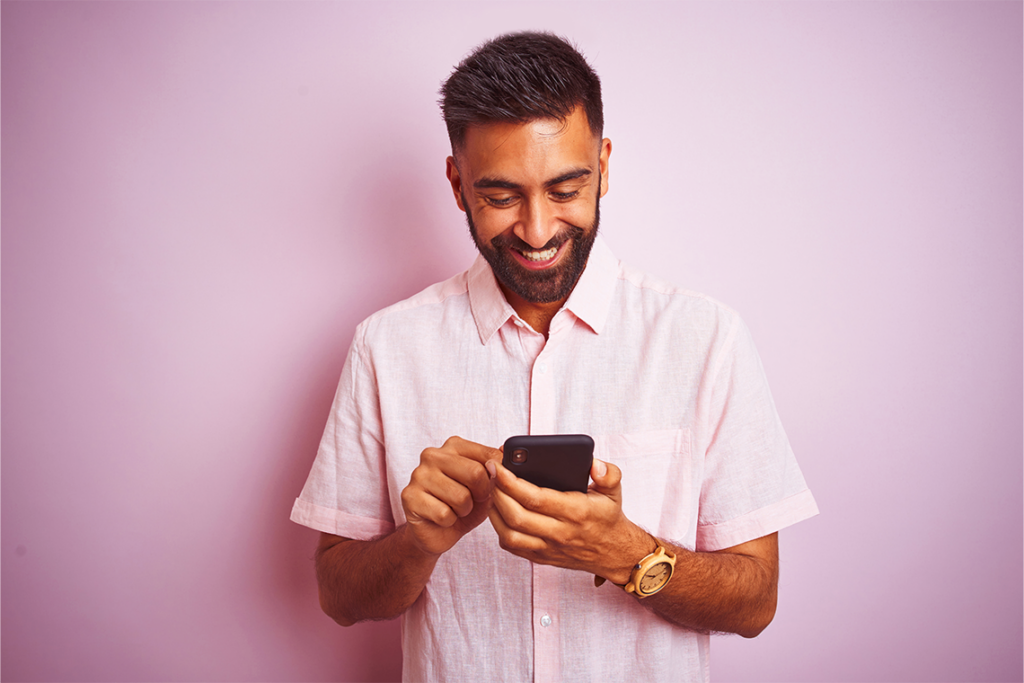 A man with a beard, wearing a pink button down shirt, smiles at his phone against a pastel purple background.